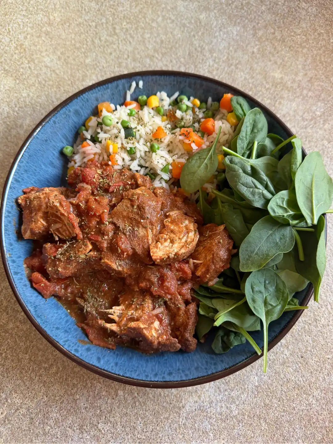 A blue bowl of chicken in a rich tomato sauce, alongside rice with peas, sweetcorn and carrot, and a handful of fresh spinach