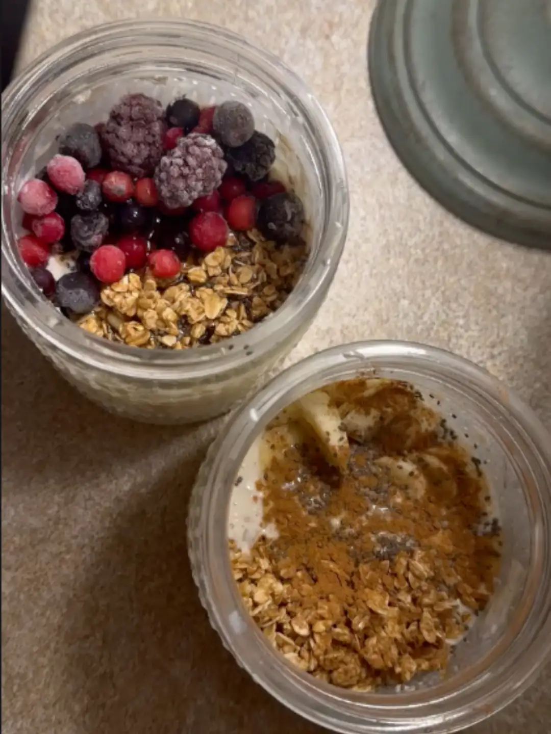Two jars of overnight oats, one with frozen berries and granola, the other with banana, cinnamon and chia