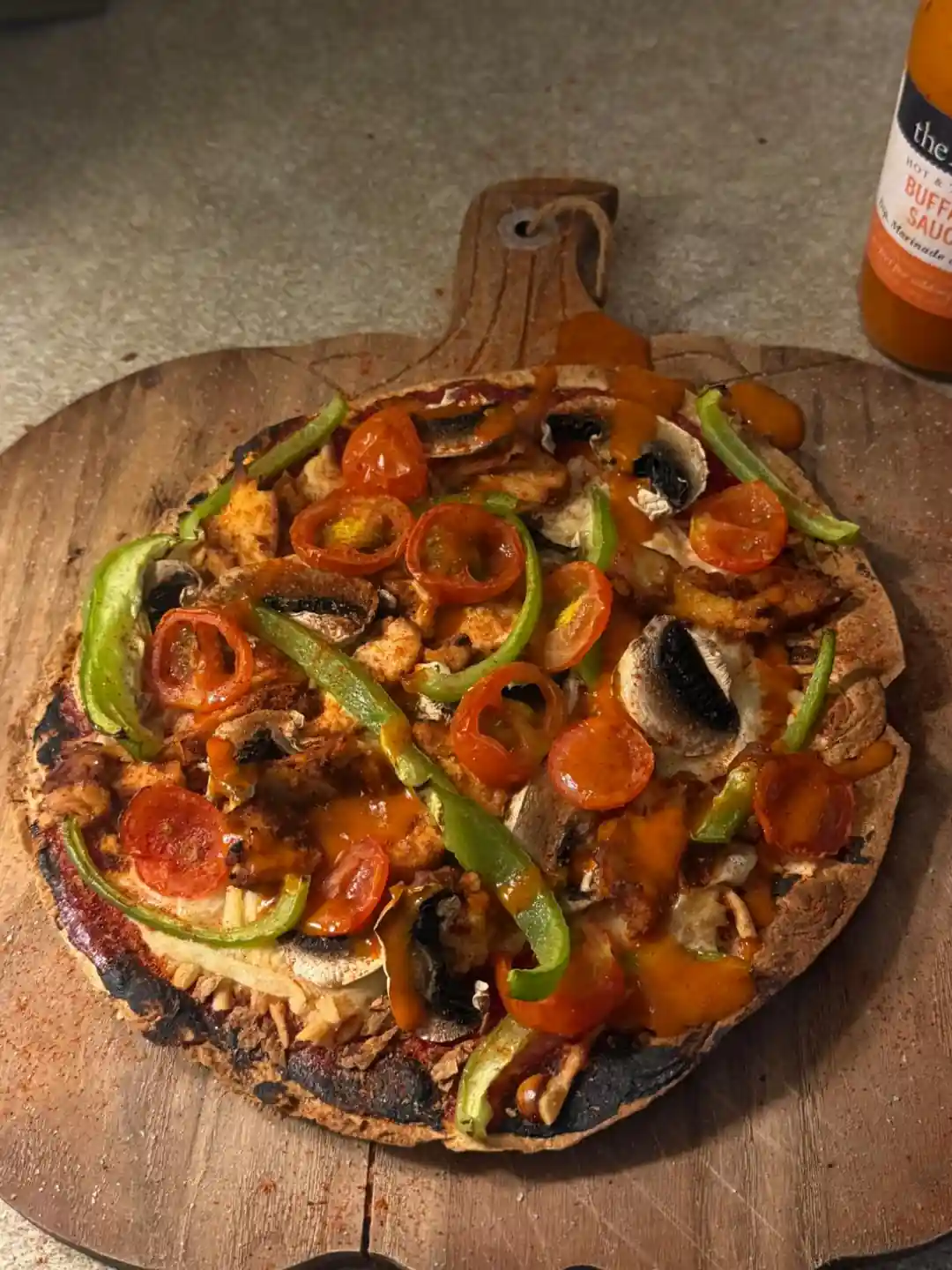 A homemade thin-crust pizza with chicken, peppers, mushrooms, tomatoes and hot sauce on a wooden board