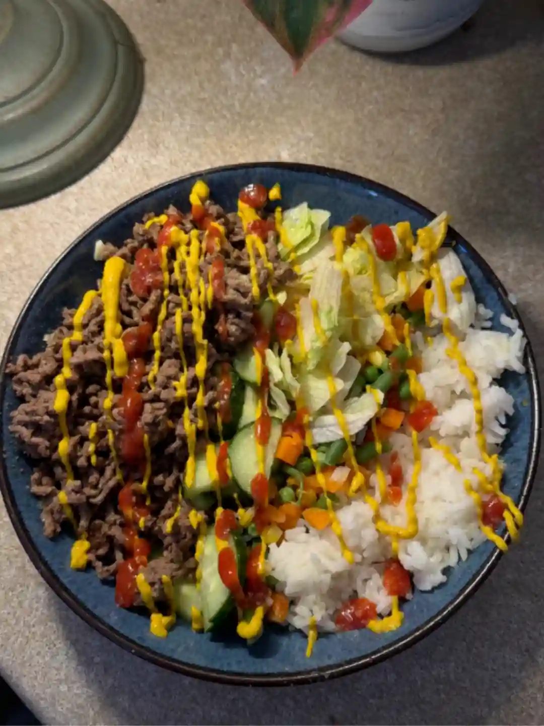 A bowl of mince, rice, cucumber and salad with mustard and hot sauce