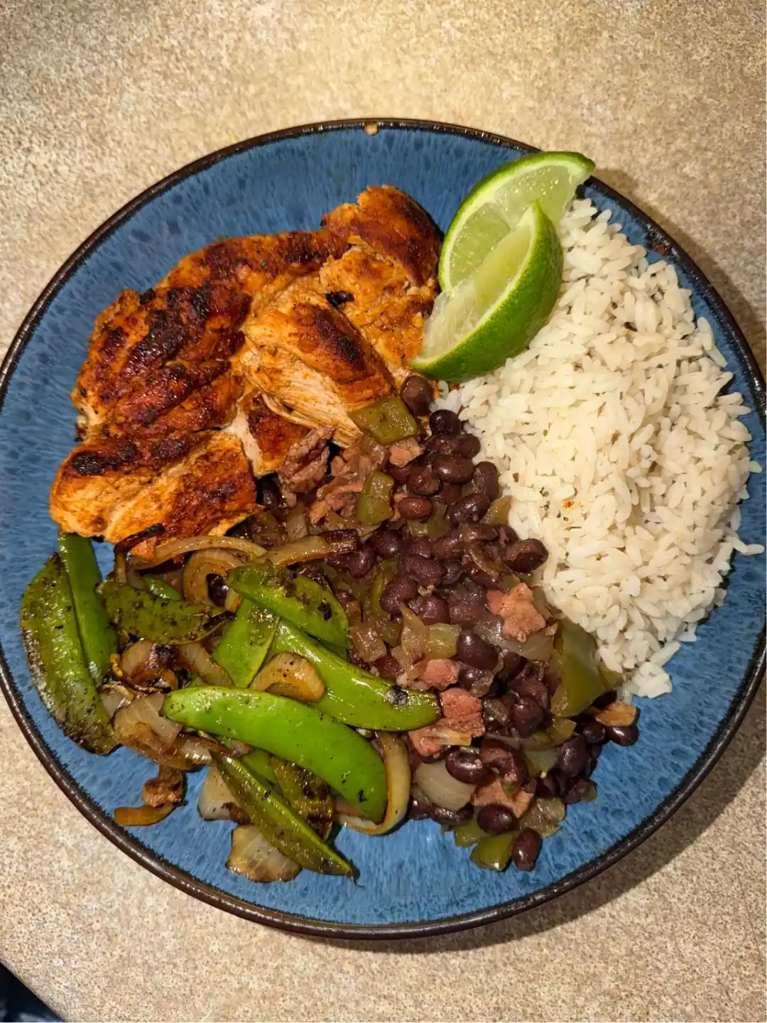 A blue bowl of sliced grilled chicken with white rice, black beans, charred peppers and onions, and two lime wedges